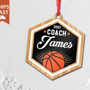 May include: A wooden hexagon-shaped ornament with a basketball design. The ornament is decorated with a black background, white trim, and a red and orange basketball. The text "2021 COACH James" is printed on the ornament.
