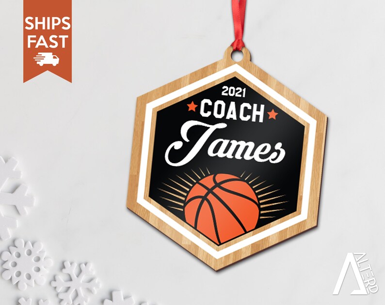 May include: A wooden hexagon-shaped ornament with a basketball design. The ornament is decorated with a black background, white trim, and a red and orange basketball. The text "2021 COACH James" is printed on the ornament.