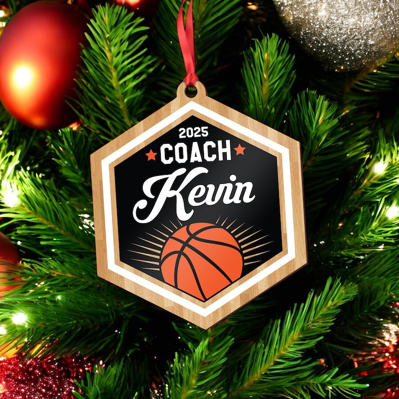 May include: A hexagonal wooden ornament with a black background and white text that reads "2025 COACH Kevin" with a star on either side of the word "COACH". A basketball with orange and black coloring is in the center of the ornament.