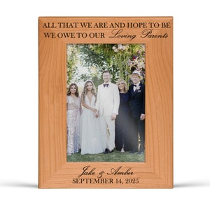 May include: A rectangular light brown wooden picture frame featuring the engraved text ALL THAT WE ARE AND HOPE TO BE WE OWE TO OUR Loving Parents at the top and Jake & Amber SEPTEMBER 14, 2025 at the bottom, holding a wedding photograph.