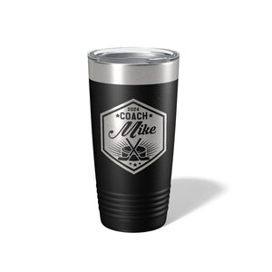 May include: A black stainless steel tumbler with a clear lid. The front features a silver laser-engraved hexagonal emblem containing the text 2024 COACH Mike, with a graphic of two crossed hockey sticks and a puck centered below the name.
