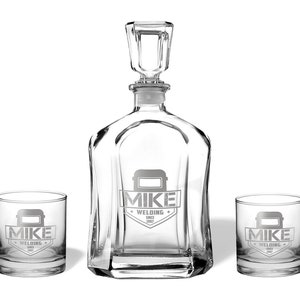 May include: A clear glass decanter set with two matching glasses. The decanter has a square stopper and a unique shape. Each piece is etched with the words "MIKE WELDING SINCE 1987" and a welding mask graphic.
