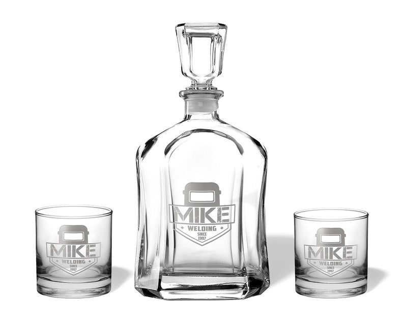 May include: A clear glass decanter set with two matching glasses. The decanter has a square stopper and a unique shape. Each piece is etched with the words "MIKE WELDING SINCE 1987" and a welding mask graphic.