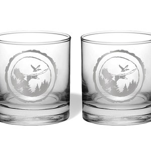 May include: Two clear glass tumblers with a design of a flying duck and trees etched into the glass. The design is in a circle that looks like a tree ring.