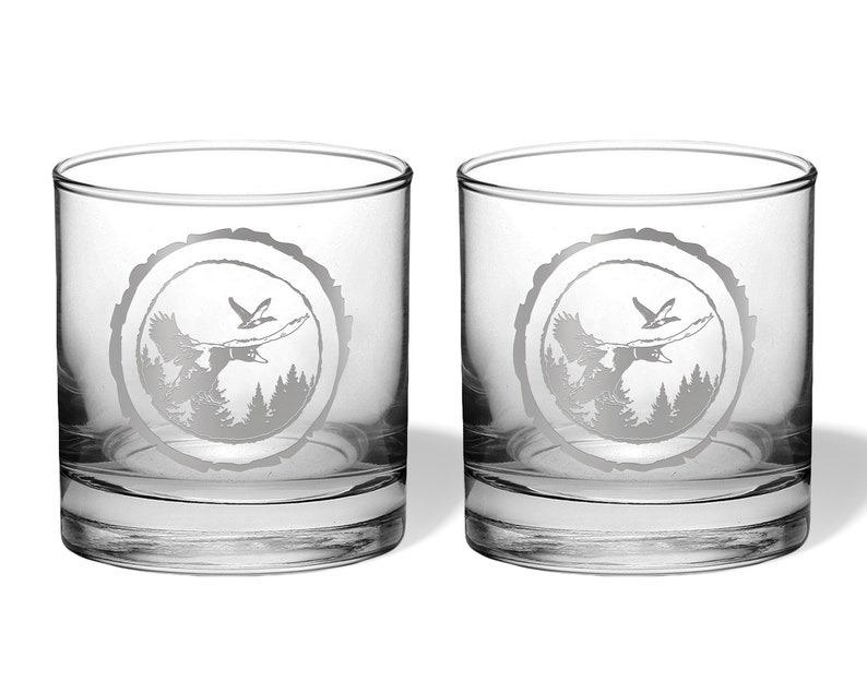 May include: Two clear glass tumblers with a design of a flying duck and trees etched into the glass. The design is in a circle that looks like a tree ring.