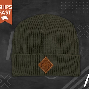 May include: A green knitted beanie with a brown leather patch featuring a mountain scene. The patch has a diamond shape and is stitched onto the front of the beanie.
