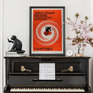 May include: A black and white poster of the movie "Vertigo" featuring James Stewart and Kim Novak. The poster is framed and hanging on a white wall above a black piano with sheet music on top.