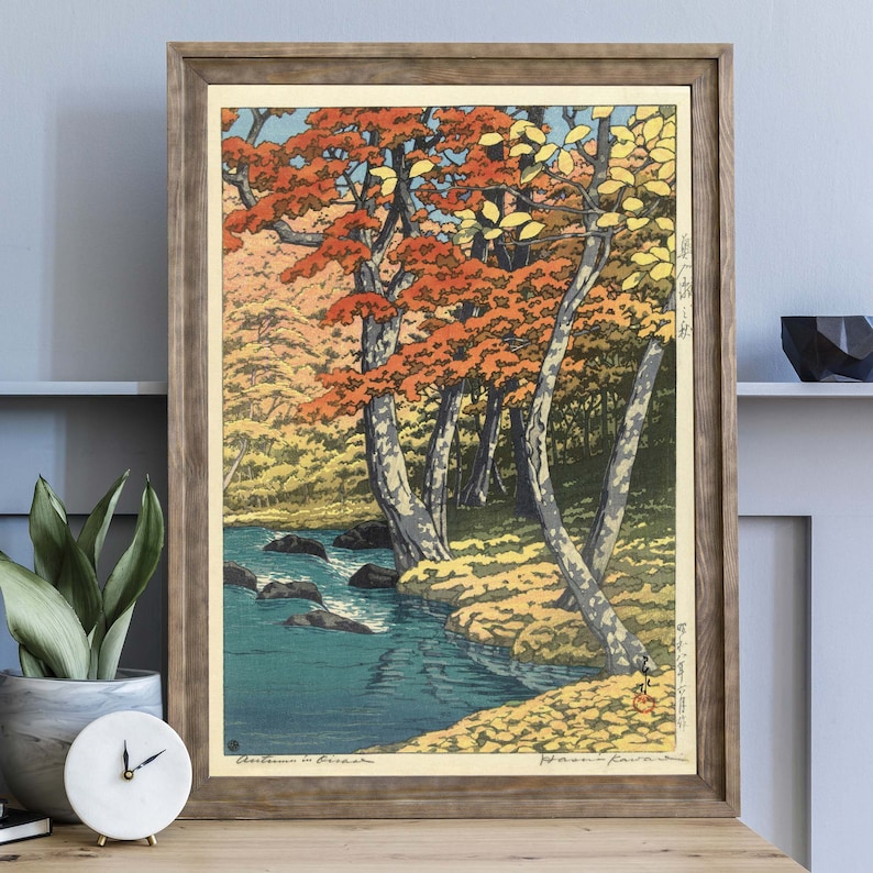 May include: A woodblock print of a Japanese landscape scene with a river flowing through a forest of trees with red, orange, and yellow leaves. The print is framed in a brown wood frame.