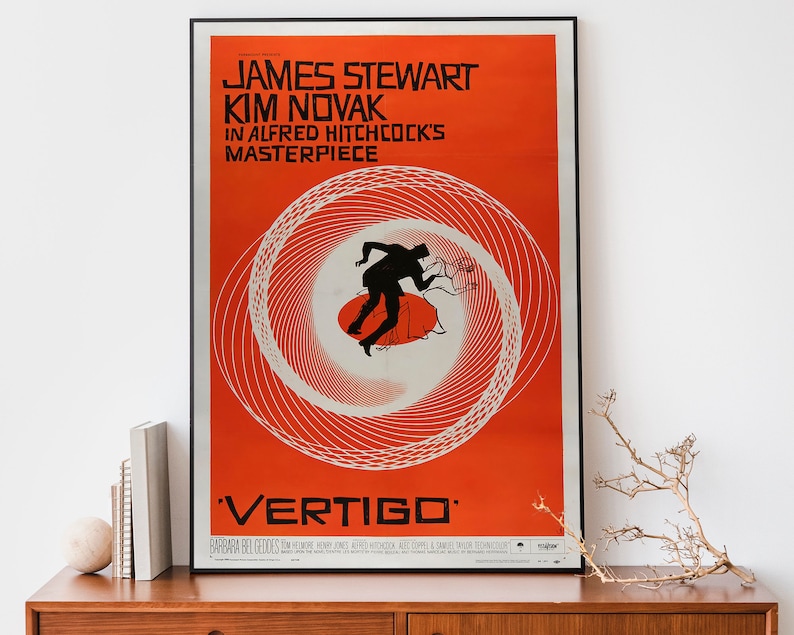 May include: A vintage movie poster for the film "Vertigo" starring James Stewart and Kim Novak. The poster features a red background with a white spiral design and a silhouette of a man falling. The text "Vertigo" is in large, white letters at the bottom of the poster.
