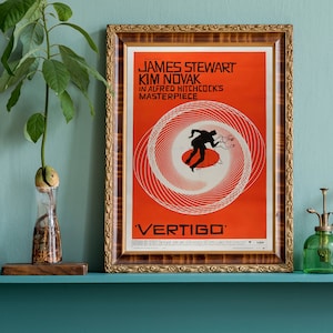 May include: A vintage movie poster for the film "Vertigo" starring James Stewart and Kim Novak. The poster features a red background with a swirling black and white design and the title "Vertigo" in large black letters.