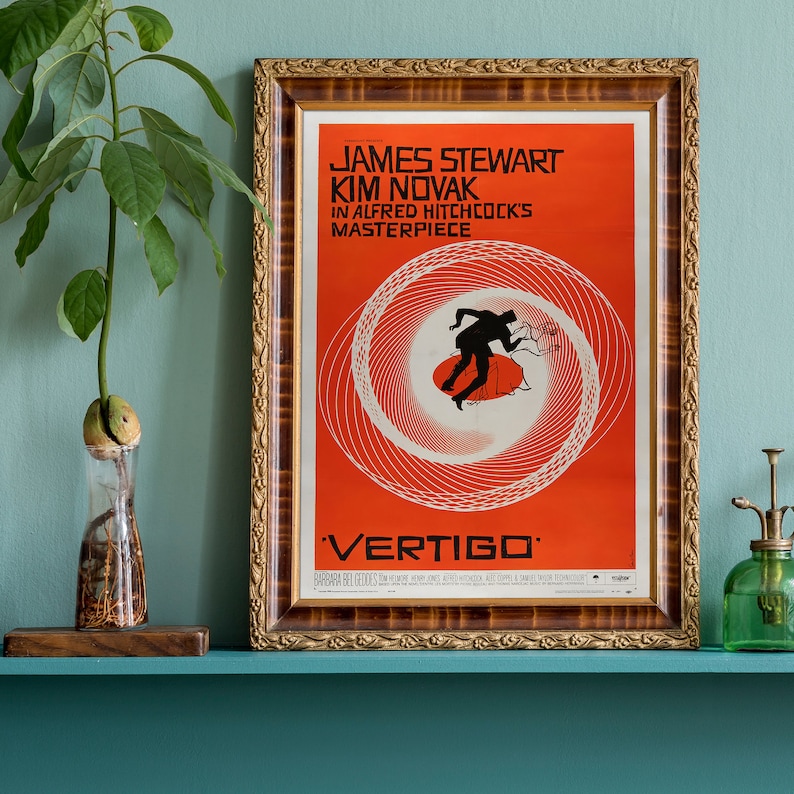 May include: A vintage movie poster for the film "Vertigo" starring James Stewart and Kim Novak. The poster features a red background with a swirling black and white design and the title "Vertigo" in large black letters.