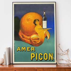 May include: Vintage poster advertising Amer Picon, a bitter orange liqueur. The poster features a woman holding a tray with a bottle of Amer Picon on it, all emerging from a large orange peel.