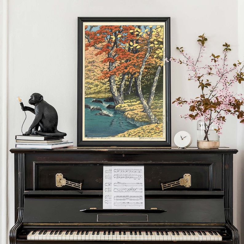 May include: A black and white woodblock print of a Japanese forest scene with a stream running through it. The print is framed in a black frame and is hanging on a white wall. There is a black monkey figurine and a clock on a black piano below the print.