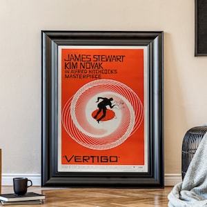 May include: A black framed movie poster with a red background and a white spiral design. The poster features the text "JAMES STEWART KIM NOVAK IN ALFRED HITCHCOCK'S MASTERPIECE 'VERTIGO'"