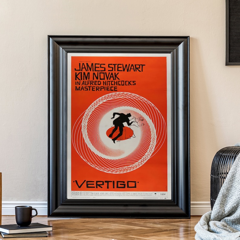 May include: A black framed movie poster with a red background and a white spiral design. The poster features the text "JAMES STEWART KIM NOVAK IN ALFRED HITCHCOCK'S MASTERPIECE 'VERTIGO'"