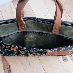 May include: A green canvas tote bag with a floral patterned front pocket. The bag has brown leather handles and a zipper closure. A small leather tag is attached to the front.