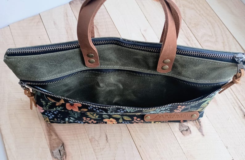 May include: A green canvas tote bag with a floral patterned front pocket. The bag has brown leather handles and a zipper closure. A small leather tag is attached to the front.