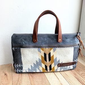 May include: A gray canvas tote bag with brown leather handles and a zipper closure. The bag features a colorful geometric pattern in shades of gray, white, yellow, and black.