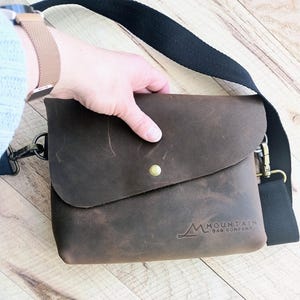 May include: A brown leather crossbody bag with a flap closure and a brass button. The bag has a black adjustable strap and the text "Mountain Bag Company" embossed on the front.