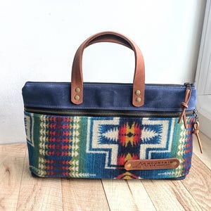 May include: A blue canvas tote bag with a brown leather handle and a zipper closure. The bag features a colorful patterned fabric lining with a geometric design. The bag has a leather tag with the text "M MOUNTAIN BAG COMPANY" on it.