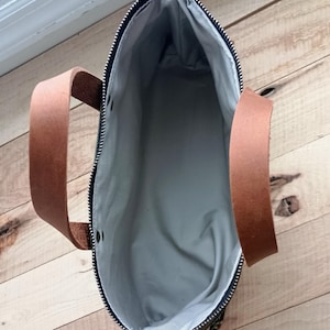 May include: An open, top-down view of a tote bag. The bag has a dark green exterior with a black zipper and brown leather handles. The interior is a light gray fabric. The bag is sitting on a wooden surface.