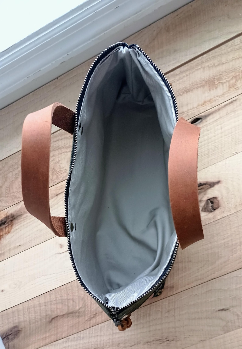 May include: An open, top-down view of a tote bag. The bag has a dark green exterior with a black zipper and brown leather handles. The interior is a light gray fabric. The bag is sitting on a wooden surface.