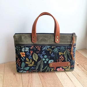 May include: A rectangular tote bag with a floral pattern on a black background. The bag has a brown leather handle and a green top. A brown leather tag with the text "MOUNTAIN BAR COMPANY" is attached to the front.