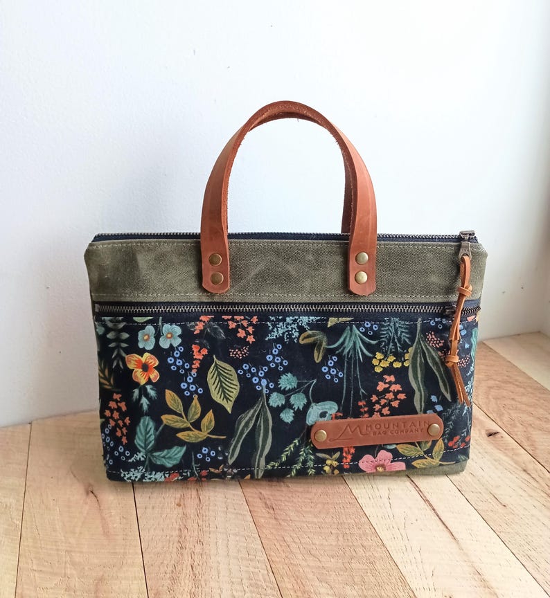 May include: A rectangular tote bag with a floral pattern on a black background. The bag has a brown leather handle and a green top. A brown leather tag with the text "MOUNTAIN BAR COMPANY" is attached to the front.