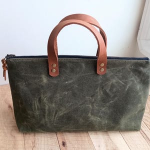 May include: An olive green tote bag with a brown leather handle. The bag has a rectangular shape with a zipper closure and a small leather pull. The handles are attached with brass rivets.