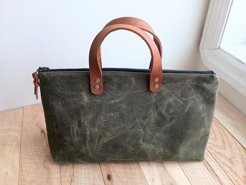 May include: An olive green tote bag with a brown leather handle. The bag has a rectangular shape with a zipper closure and a small leather pull. The handles are attached with brass rivets.