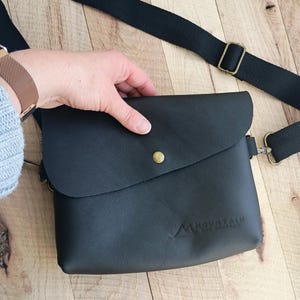 May include: Black leather crossbody bag with a flap closure and a gold button. The bag has a logo that says "Mountain Supply" on the front.