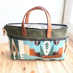 May include: A green and blue patterned tote bag with leather handles. The bag has a zipper closure and a small leather tag that says "Mountain Made Co."