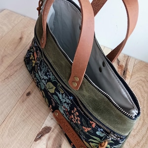 May include: A floral print handbag with olive green accents and brown leather handles. The bag features a black background with colorful flowers and foliage. The handles are attached with metal rivets.