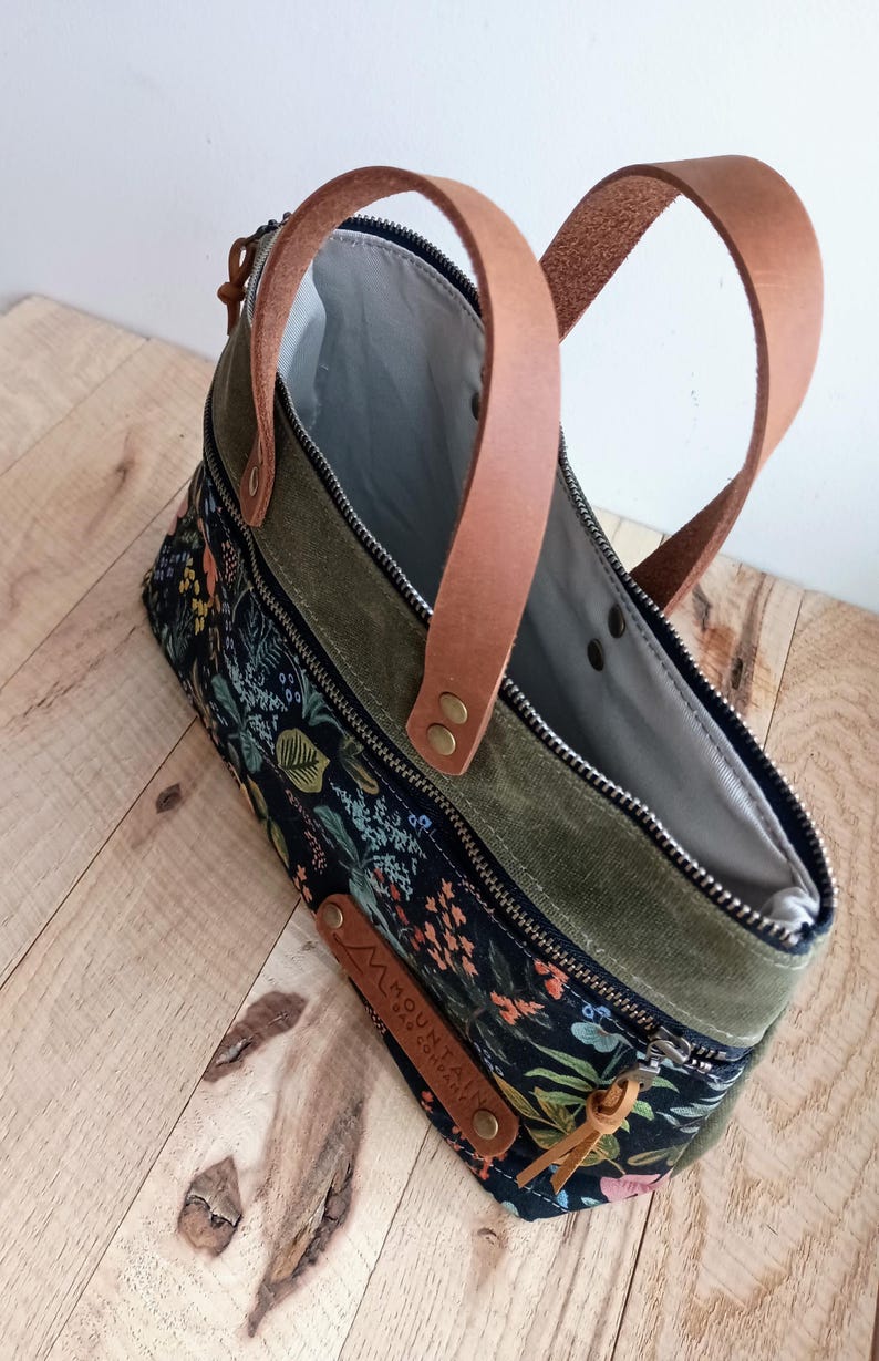 May include: A floral print handbag with olive green accents and brown leather handles. The bag features a black background with colorful flowers and foliage. The handles are attached with metal rivets.