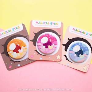 Può includere: Tre spille rotonde colorate con un design a occhio da cartone animato. Ogni spilla presenta uno schema di colori e un design diversi, tra cui rosa, blu e arancione. Le spille sono etichettate "Magical Eyes".