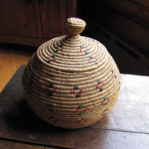 May include: A round, woven basket with a lid. The basket is made of natural-colored fibers and features a spiral design. Small squares of red, blue, and green are woven into the basket. The lid has a small, rounded knob on top. The basket is sitting on a wooden surface.