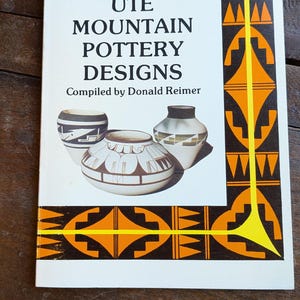 May include: The cover of a book titled "UTE MOUNTAIN POTTERY DESIGNS" with illustrations of pottery. The cover has a white background with orange and black geometric designs. The book is compiled by Donald Reimer.