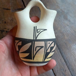 May include: A small, cream-colored ceramic vessel with a bridge handle and two openings. The lower section features a horizontal band decorated with black geometric line patterns and subtle tan shading against the matte surface.
