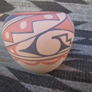 May include: A hand-painted ceramic bowl with a rounded shape. The bowl features a cream-colored base with geometric designs in red and dark blue. The design includes zigzags, curves, and dotted patterns. The rim is also painted red.