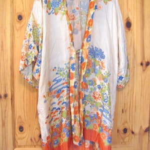 May include: A vintage kimono-style robe in cream with a floral pattern in orange, blue, and green. The robe has wide sleeves and a tie closure. The hem and trim are a vibrant orange.