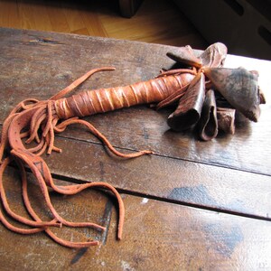 May include: A handcrafted, rustic leather rattle with a long, wrapped handle and a cluster of bell-shaped elements at the top. The rattle features a warm, earthy color palette of brown and tan, with long, dangling leather fringe.