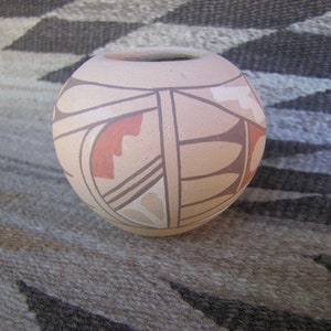 May include: A round, beige pottery vase with a small opening at the top. The vase is decorated with geometric patterns in shades of brown, red, and cream. The design features lines, shapes, and stylized motifs, creating a Southwestern aesthetic.