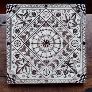 Puede incluir: Azulejo cerámico cuadrado con un diseño detallado de flores y hojas en tonos marrones y blancos. El azulejo presenta un patrón circular central rodeado de hojas estilizadas, flores y bordes geométricos.