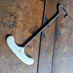 May include: An antique, rusty metal hook with a white handle. The handle is curved and has dark lines. The hook is on a wooden surface with a dark brown color.