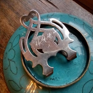 May include: A silver metal trivet with an ornate design, resting on a teal-colored dish with a floral pattern. The trivet has a heart-shaped top and a monogram. The dish has a gold rim.