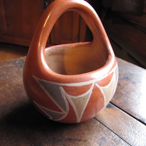 May include: A handmade, terracotta-colored ceramic basket with a handle. The basket features a geometric design in white, gray, and tan around the rim. The basket is sitting on a wooden surface.