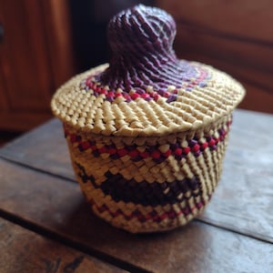 May include: A woven basket with a lid. The basket is made of natural-colored straw with red and purple accents. The lid has a rounded top and is also woven. The basket is sitting on a wooden surface.