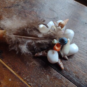 May include: A handcrafted wand featuring a wooden handle, adorned with a cluster of seashells, feathers, and a small, colorful shell. The wand's design incorporates various shell shapes and colors, creating a unique, natural aesthetic.