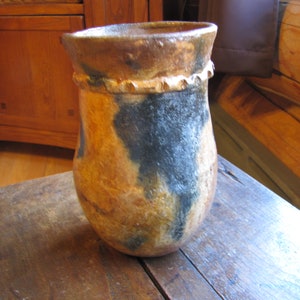 May include: A handcrafted ceramic vase with a unique, organic shape. The vase features a blend of earthy tones, including brown, tan, and black, with a textured surface. The rim has a decorative, scalloped edge, adding a rustic charm to this decorative piece.