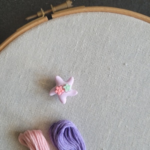 Op de afbeelding: Een lichtpaarse stervormige versiering met twee kleine bloemaccenten, geplaatst op een linnen stof die is gespannen in een houten borduurring. Twee strengen borduurgaren, één roze en één paars, zijn ook zichtbaar.